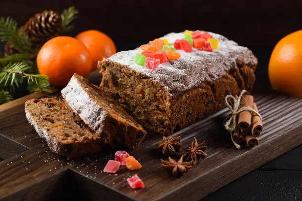 Traditional sliced freshly baked fruit cake decorated with candied fruits, star anise, cinnamon and clementines on dark background closeup
