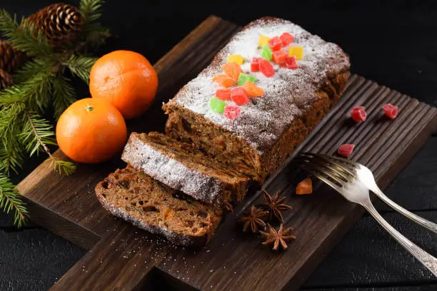 Delicious homemade fruit cake decorated with sugar icing and candied fruits served with clementines and fir branch on oak board closeup