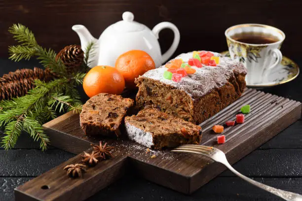 Traditional English fruit cake with candied fruits served with black tea, clementines and fir branch on oak board with silverware