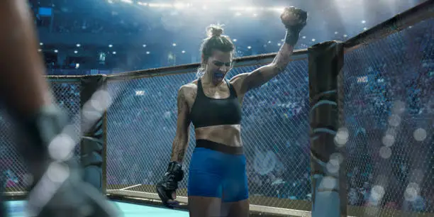 A close up composite image of a professional female mixed martial arts fighter dressed in tight shorts, sports bra, and grappling gloves. The fighter is wearing a mouth guard and raising her fist whilst screaming in victory. She stands in an octagon cage in a generic indoor sports arena.
