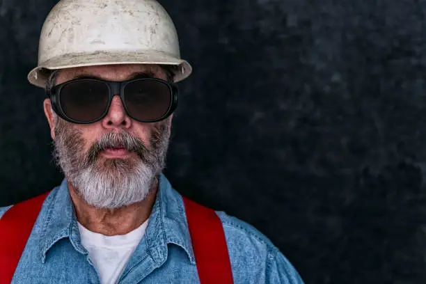 Portrait of a  middle-aged Caucasian construction worker wearing safety-glasses looking at the camera. He is also wearing a worn white hard hat, blue denim shirt, red suspenders and a white t-shirt. He has an unkempt graying beard.