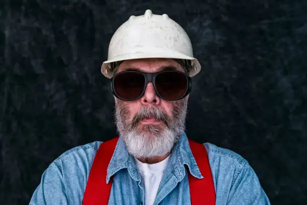 Portrait of a  middle-aged Caucasian construction worker wearing safety-glasses looking at the camera. He is also wearing a worn white hard hat, blue denim shirt, red suspenders and a white t-shirt. He has an unkempt graying beard.