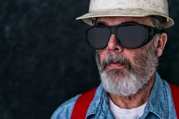 Profile portrait of a  middle-aged Caucasian construction worker wearing safety-glasses looking off. He is also wearing a worn white hard hat, blue denim shirt, red suspenders and a white t-shirt. He has an unkempt graying beard.