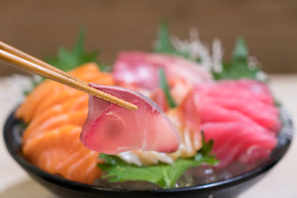 Chopsticks with Hamachi sashimi with Mixed sliced fish sashimi on ice in black bowl. Sashimi Salmon Tuna Hamachi Prawn and Surf Calm set, raw fish, japanese food in Asian restaurant.