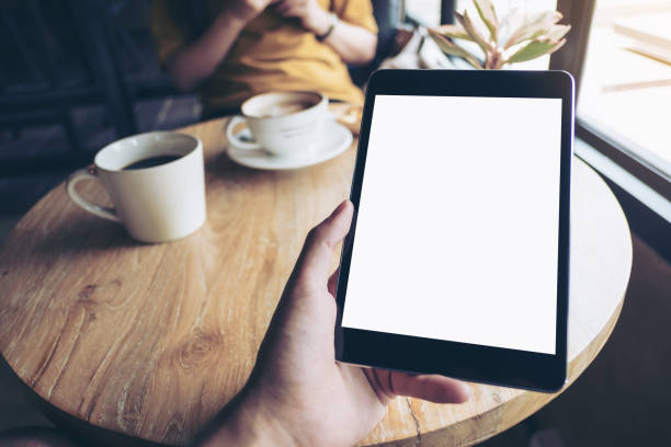 istock Mock up image of a hand holding black tablet pc with white blank screen on wooden table with woman using mobile phone in background in modern cafe 913599302