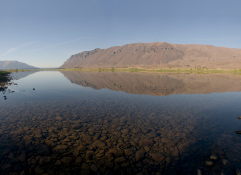 Early morning during arctic sunrise. Mountain with a snow and its reflection in the water of a fjord. Blue sky with light clouds. Sun still low. Nordaustur, North-West Iceland.