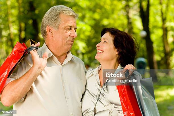 Foto de Feliz Casal Senior Sorridente Com Sacos De Compras Caminhando No Parque e mais fotos de stock de Abraçar