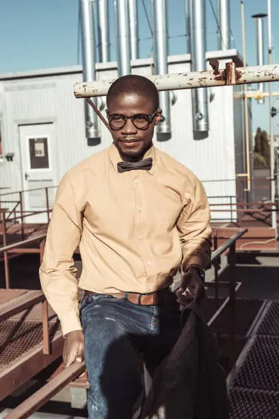Stylish african american man wearing glasses and bowtie, sitting on guardrails and looking aside