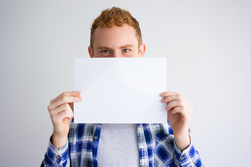 Smiling red haired Caucasian guy covering lower face with empty sheet of paper. Student presenting something on paper. Advertising concept
