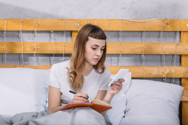 istock girl writing something to notebook and looking at smartphone in bed 913718034