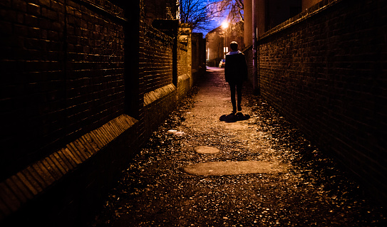 A young man walking home alone at night through a dark alleyway in the UK.