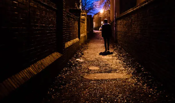 A young man walking home alone at night through a dark alleyway in the UK.