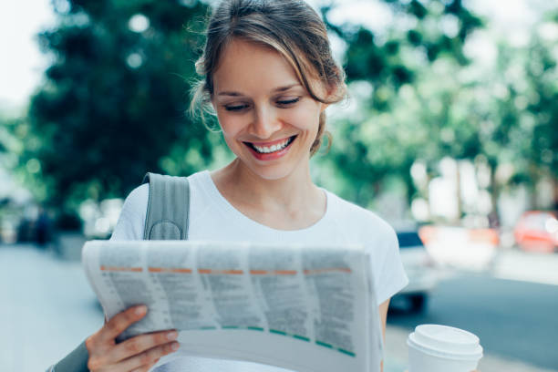 Smiling Woman Reading Newspaper on Street Closeup portrait of smiling young beautiful woman holding drink and reading newspaper on street. Front view. reading newspaper stock pictures, royalty-free photos & images