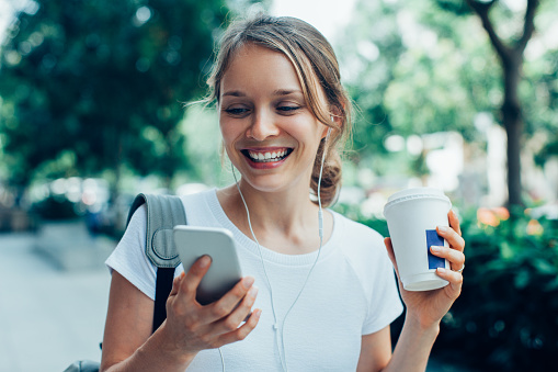 Closeup portrait of smiling young beautiful woman walking, holding drink, wearing headphones and having video call on smartphone on street