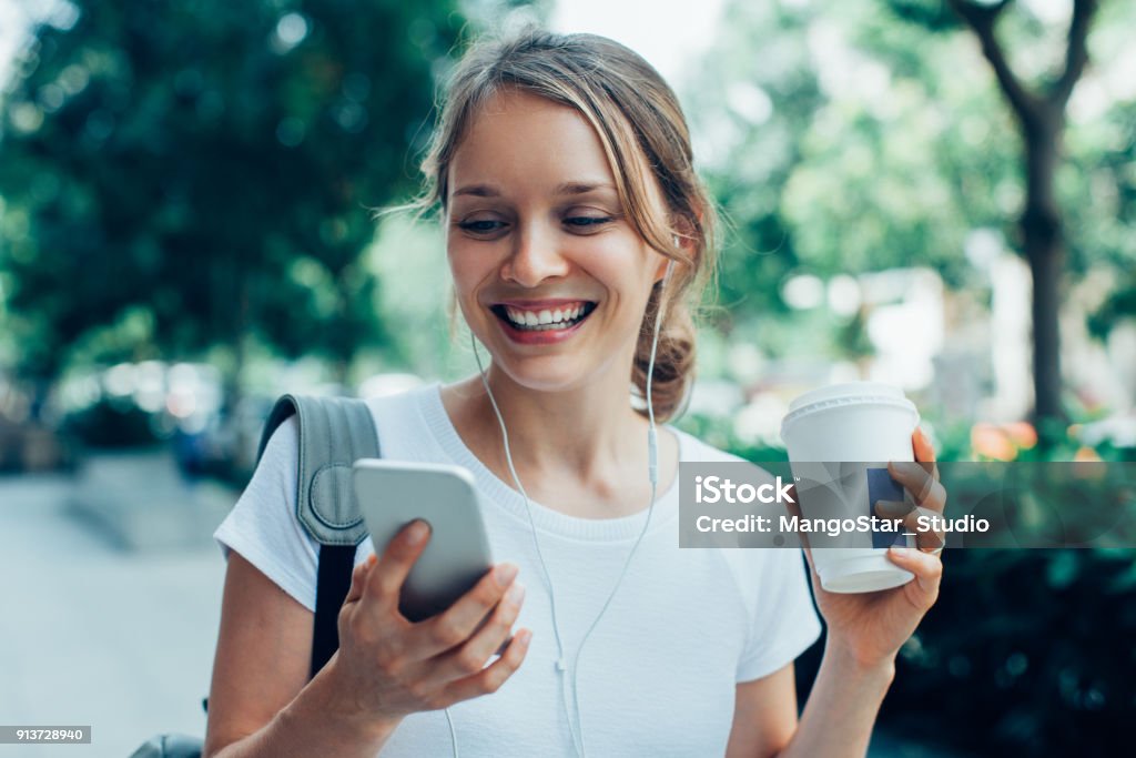 Smiling Young Woman Having Video Call Outdoors Closeup portrait of smiling young beautiful woman walking, holding drink, wearing headphones and having video call on smartphone on street Women Stock Photo