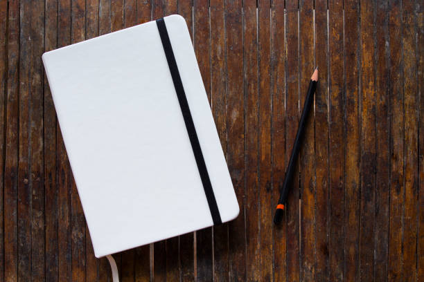 White cover notebook with black pencil on rustic wooden table flat lay photo. White cover notebook with black pencil on rustic wooden table flat lay photo. Closed notebook with blank cover flat lay photo. Notepad on table top view. Sketchbook banner template. Art logo mockup moleskin stock pictures, royalty-free photos & images