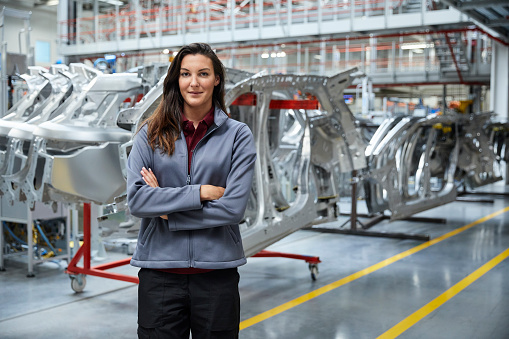 Portrait of female engineer standing against car chassis. Confident technologist is smiling in automobile industry. She is with arms crossed in factory.