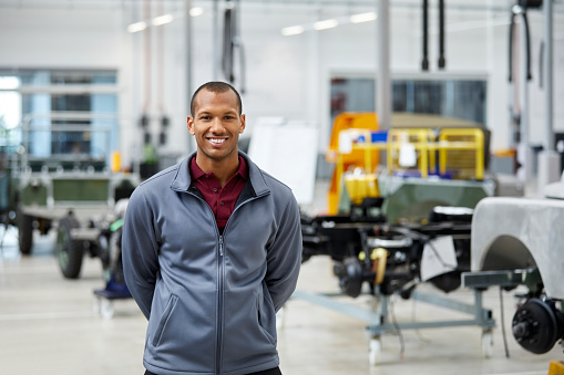 Portrait of engineer smiling in automobile industry. Confident male technologist is standing in showroom. He is wearing jacket in factory.