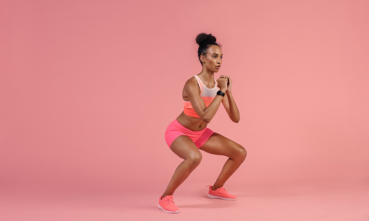 Sporty woman practicing squat exercises in studio. African woman in sportswear working out on pink background.