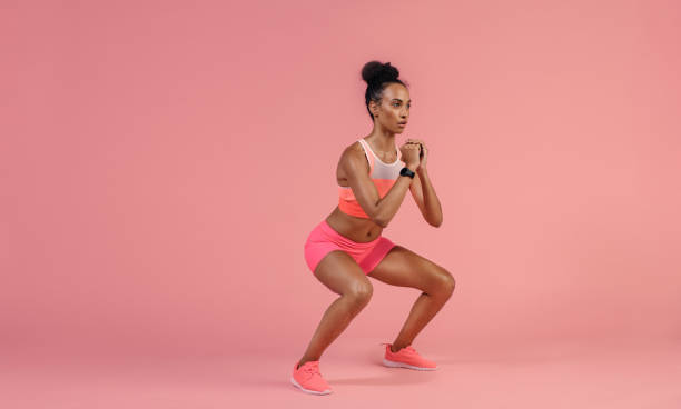Sporty woman practicing squat exercise Sporty woman practicing squat exercises in studio. African woman in sportswear working out on pink background. squatting position photos stock pictures, royalty-free photos & images