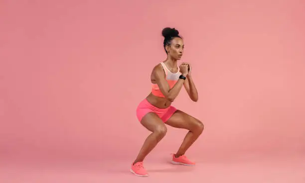 Sporty woman practicing squat exercises in studio. African woman in sportswear working out on pink background.
