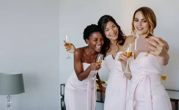 Three bridesmaids taking selfie in a room. Smiling young women having drinks and taking selfie with mobile phone.