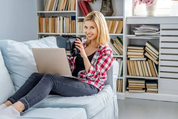 Blond female sitting on a sofa and using laptop with virtual reality glasses device.