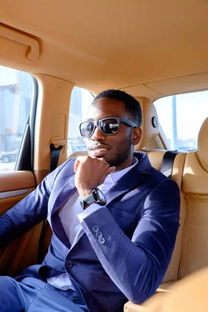 Black man in a suit and sunglasses sitting in a car.