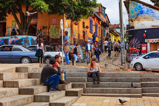 Bogotá, Colombia - Local Colombians And Tourists In The Historic La Candelaria District of The Andean Capital City. Background: Calle Del Embudo. Bogota, Colombia - May 28, 2017: A few tourists and and many local Colombians in the historic La Candelaria District of the Capital city. In the background is the narrow Calle del Embudo which gets its name from its shape. The English translation of the name would be, "Funnel Street." It is one of the most colorful streets in the capital city of the South American country of Colombia. Constructed over 450 years ago, the street leads to the Chorro de Quevedo, the plaza where it is believed the Spanish Conquistador, Gonzalo Jiménez de Quesada founded the city in 1538. Many street facing walls in this area are painted with either street art or the legends of the pre-Colombian era, in the vibrant colours of Colombia. Photo shot in the afternoon sunlight; horizontal format. Camera: Canon EOS 5D MII. Lens EF 24-70 mm F2.8L USM. calle del embudo stock pictures, royalty-free photos & images
