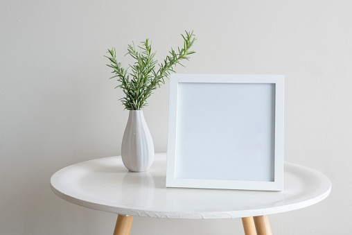 Close up of sprigs of rosemary in small white vase with blank square frame on round table against beige wall