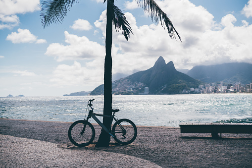 Silhouettes of abandoned bicycle tied to a palm tree and wooden bench on pavement stone; view from Arpoador beach in Rio de Janeiro with the Two Brother hills and warm carioca sea in the background