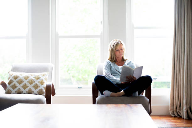 mulher madura, lendo o livro na cadeira em casa - women book mature adult reading - fotografias e filmes do acervo