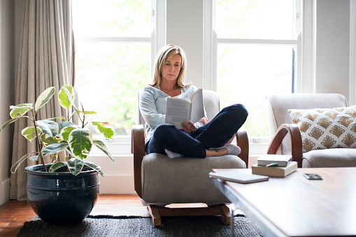 Full length of mature woman reading book on chair. Beautiful female is relaxing by houseplant against window. She is wearing casuals at home.