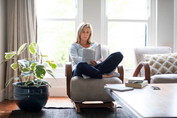 Mature woman reading book on chair at home Full length of mature woman reading book on chair. Beautiful female is relaxing by houseplant against window. She is wearing casuals at home. woman sitting cross legged in chair stock pictures, royalty-free photos & images