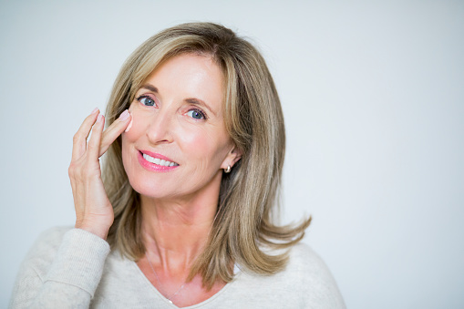 Portrait of mature woman applying moisturizing cream. Beautiful female is taking care of her skin. She is with blond hair against white background.