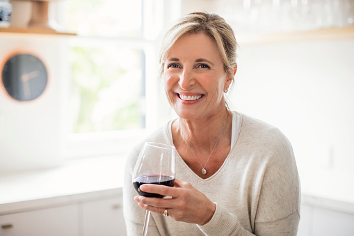 Portrait of smiling mature woman holding wineglass in kitchen. Beautiful female is enjoying wine at home. She is wearing casuals.