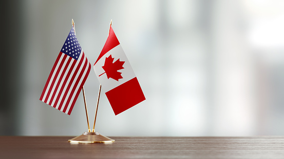 American and Canadian flag pair on desk over defocused background. Horizontal composition with copy space and selective focus.