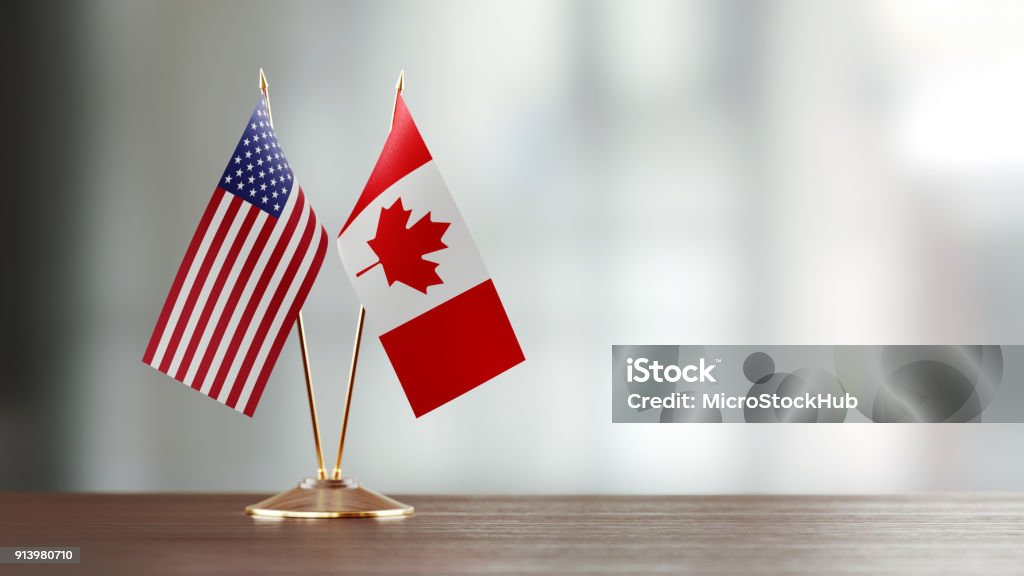 American And Canadian Flag Pair On A Desk Over Defocused Background American and Canadian flag pair on desk over defocused background. Horizontal composition with copy space and selective focus. Canada Stock Photo