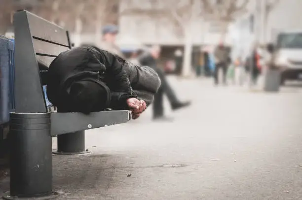 Photo of Poor homeless man or refugee sleeping on the wooden bench on the urban street in the city, social documentary concept, selective focus
