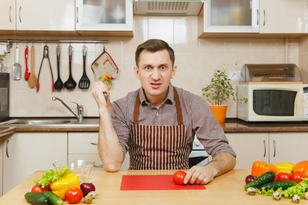 istock Angry crazy caucasian young man in apron, brown shirt sitting at table, attack cuts vegetable for salad with knife in light kitchen. Dieting concept. Healthy lifestyle. Cooking at home. Prepare food. 914007040