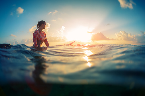 Young lady surfer waits the waves in ocean during sunrise with surf board.