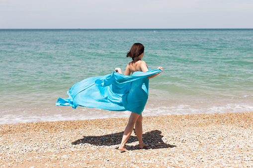 Girl in blue pareo on beach. Woman in blue sarong walks with arms outstretched along seashore