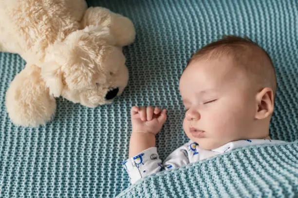 Photo of sleeping newborn baby on a blanket with a teddy. Peaceful baby lying on a bed while sleeping in a bright room with his teddy bear. My Best Friend.  Family at home. Love. Baby newborn portrait, boy kid newborn resting with toy dog friend.