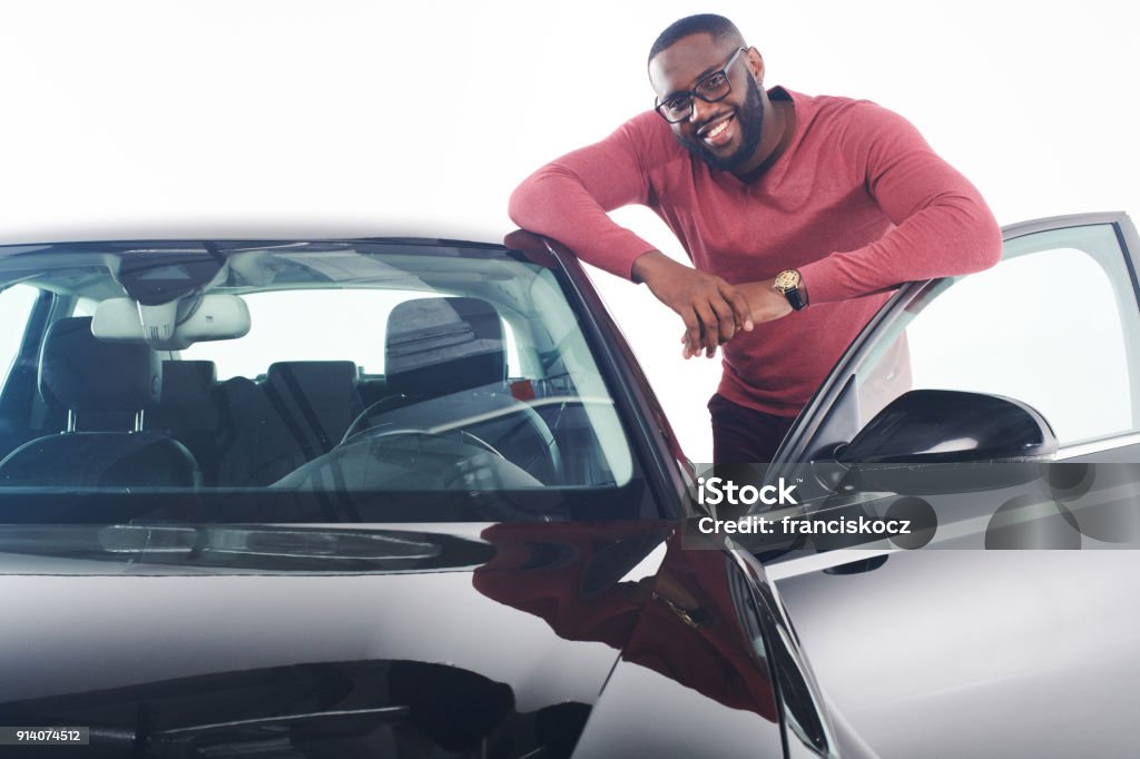 Mi sento orgoglioso. Attraente giovane africano in abito elegante in piedi vicino all'auto di lusso nera modello 2018 e appoggiato sul tetto. - Foto stock royalty-free di Automobile