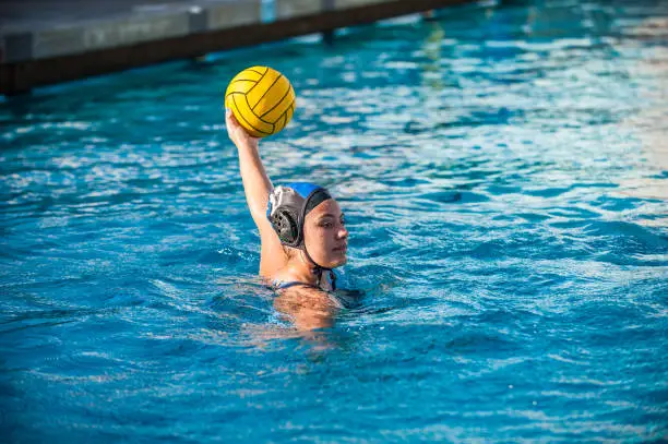 High School female water polo player holding ball high above the water looking to pass or shoot.