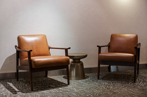 Two empty retro beige leather arm chairs in the middle of hotel hall sitting on old carpet.