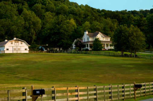 Photo is of a Farm in the Country with White Barn and White House or Home. the yard is green grass or lawn with a wooden fence around the entire property. there are mailboxes in front of the fence. and there are woods made of trees surrounding the entire property. the grass and leaves are green during the summer or spring. and the sky is blue. and the lighting is natural sunlight. 