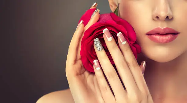 Young appealing woman is holding red rose and showing pale-pink french style manicure with  rhinestone on the slender fingers. Perfect makeup and trendy manicure.