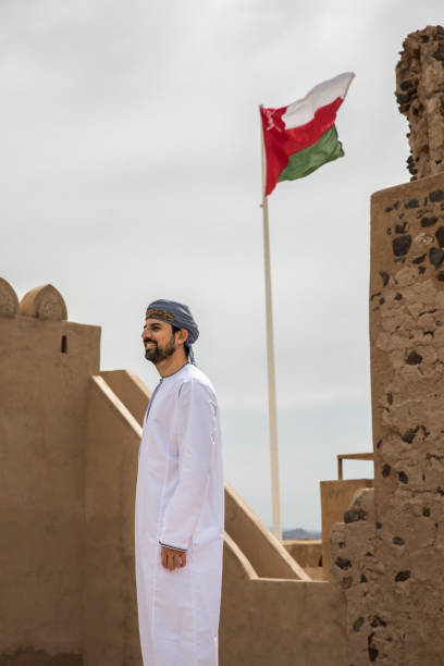 arab man in traditional omani outfit with omani flag flying at the background stock photo