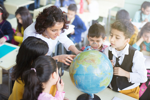female teacher and students looking the globe with a magnifying glass in the classroom